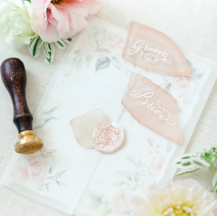 Flat lay of a floral wedding invitation suite with vellum jacket, pink sea glass place cards labeled Bride and Groom in white calligraphy, a petal wax seal, and a wax seal stamp from a styled shoot.