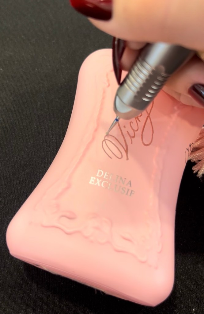 Close-up of Jessica Wofford engraving the name Vicky onto a small light pink Parfums de Marly perfume bottle during Nordstrom’s All-In personalization event in the Houston Galleria.