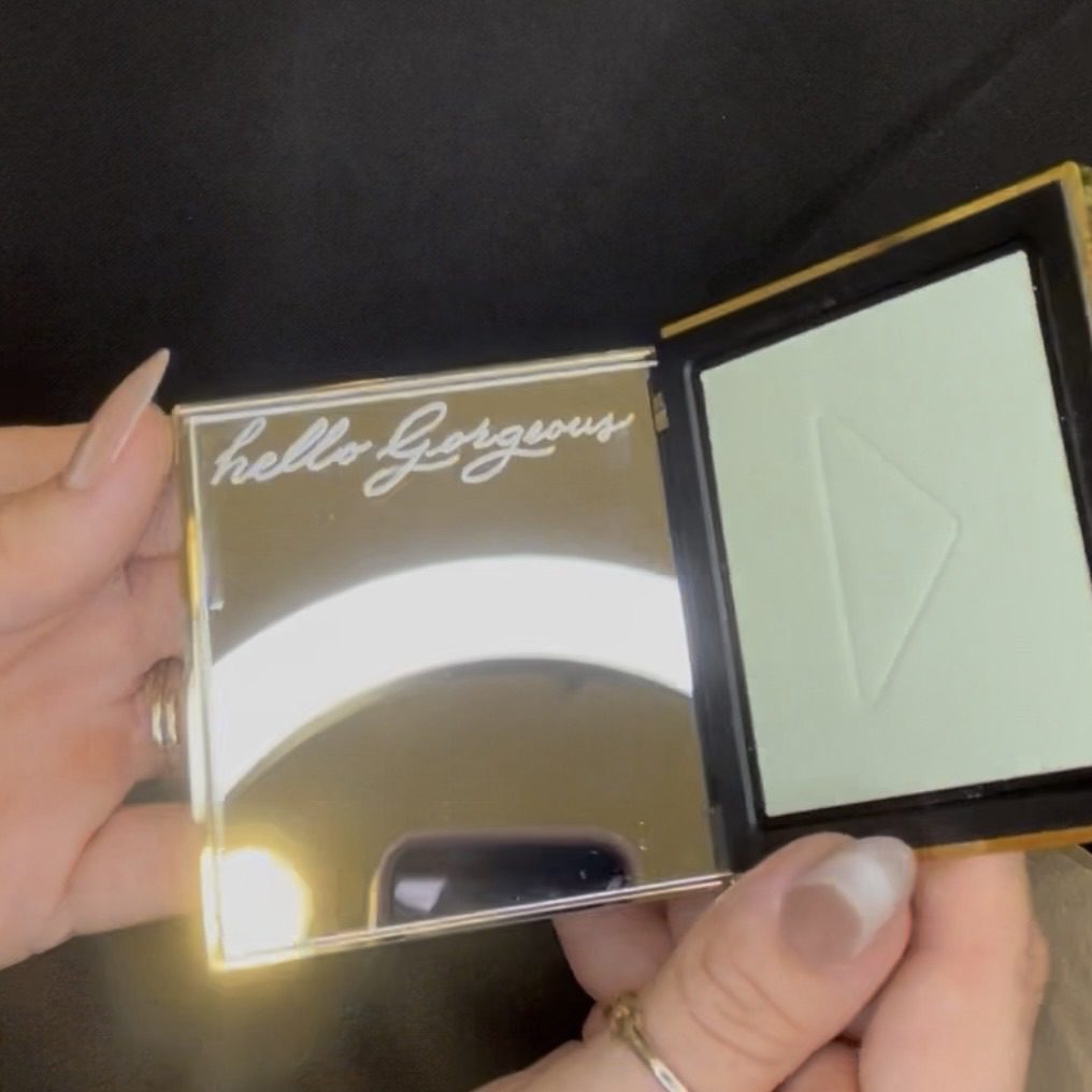 Close-up of a Prada gold compact mirror being held open, showing the engraved phrase “Hello Gorgeous” on the mirror surface.