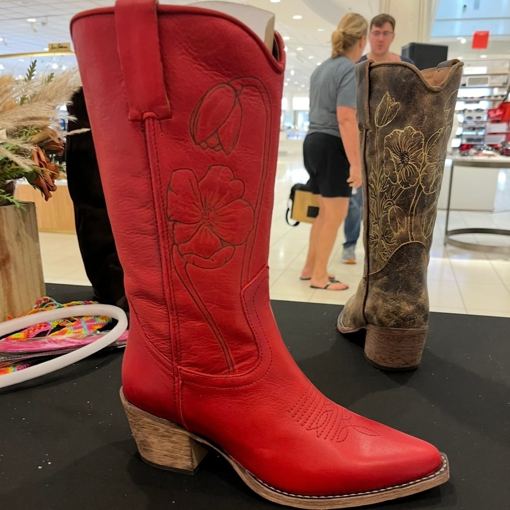 Bright red Steve Madden boot featuring a hand-burned floral design on the top front sleeve, with a brown Steve Madden boot in the background customized with mixed-media floral artwork including burned lines and gold paint accents, created during the Steve Madden boot-burning and hat bar activation at Nordstrom in the Houston Galleria.