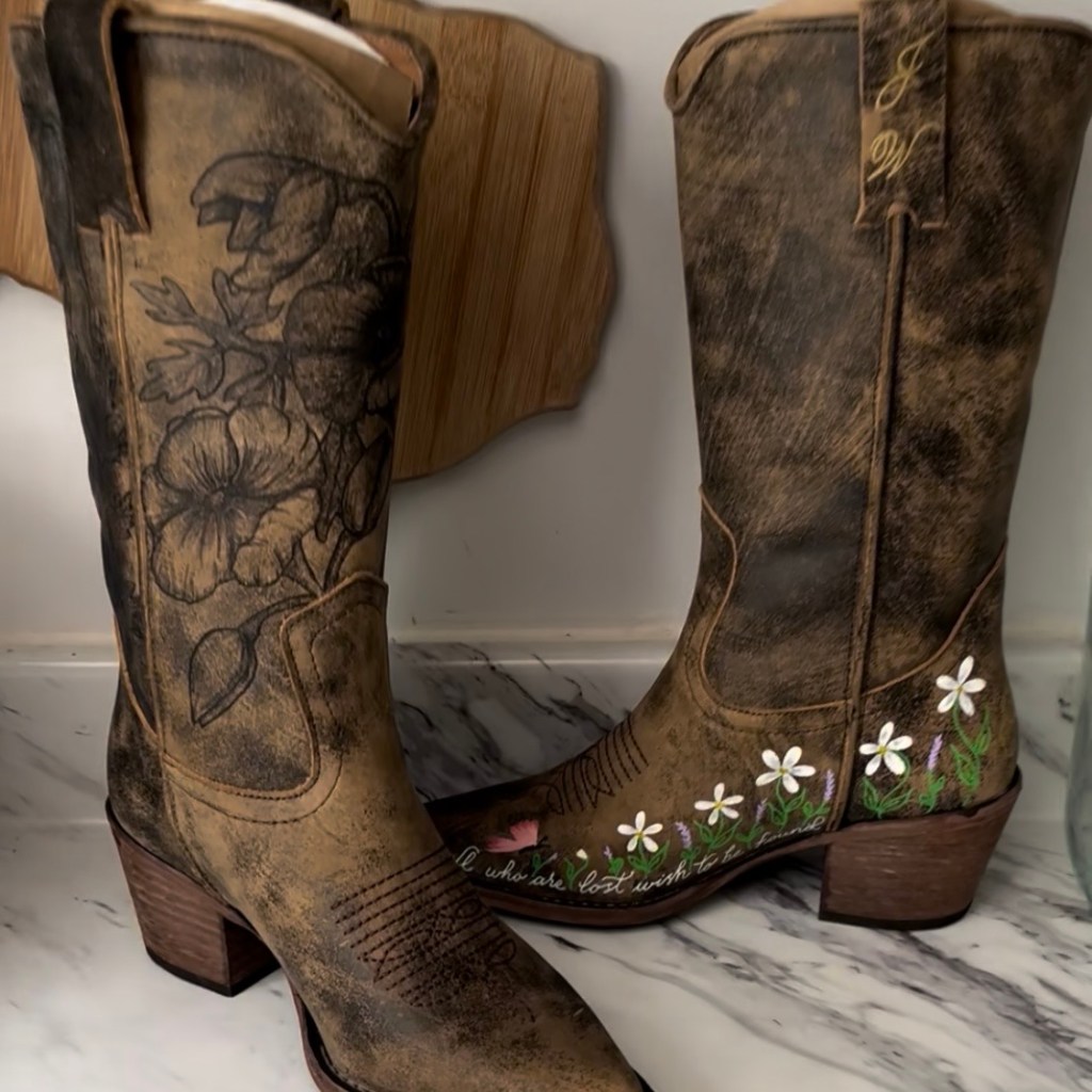 Two Steve Madden western-style boots customized with floral designs, one featuring hand-burned leather engraving and the other painted with white and green flowers, created by Houston calligrapher and engraver Jessica Wofford.