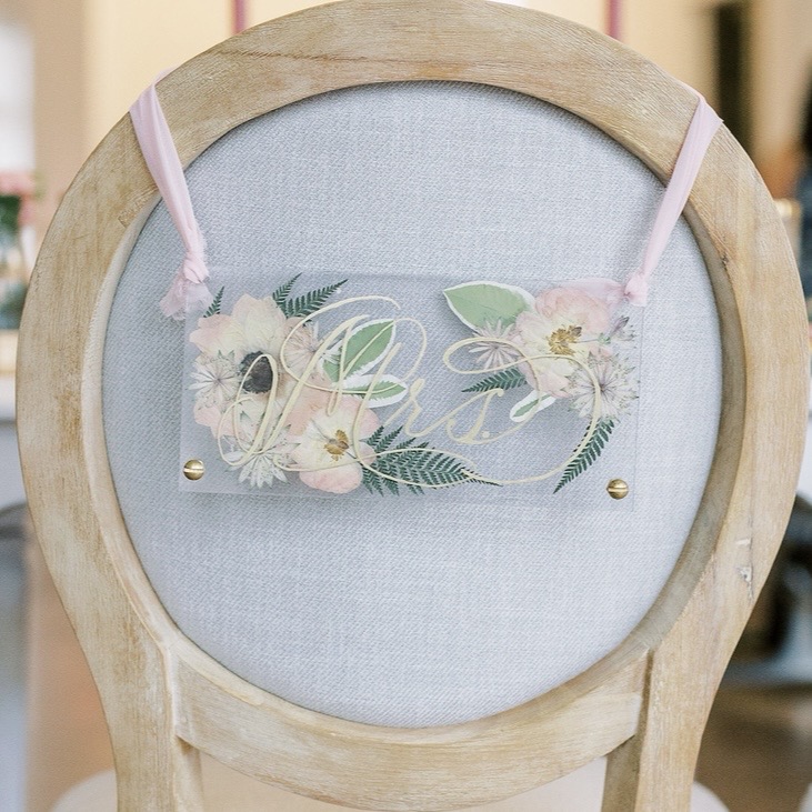 Acrylic pressed flower chair hanger with gold calligraphy reading Mrs., attached to a wooden chair with pink ribbon at a styled shoot.