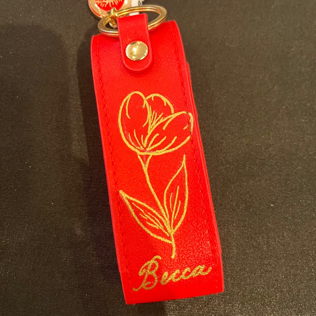 Red lipstick keychain case hand painted with a gold floral design and the name Becca during an Omorovicza live personalization event