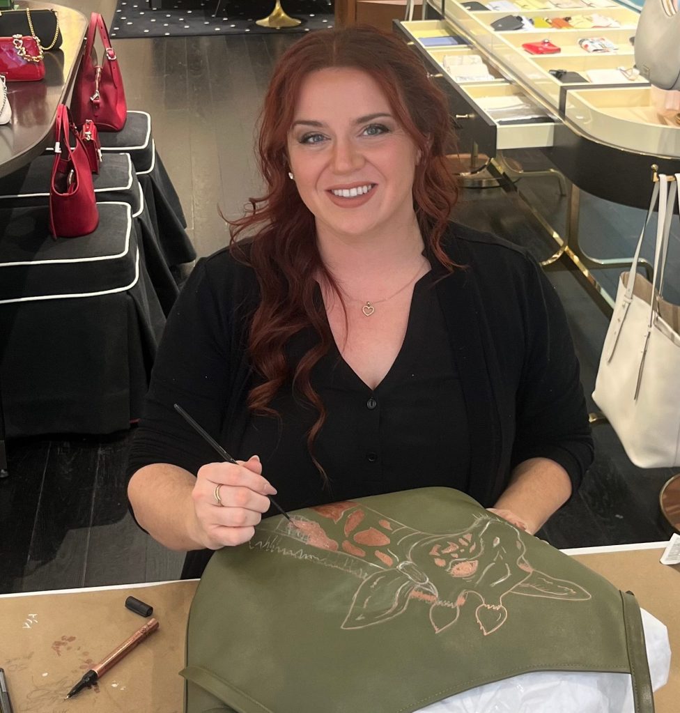 Houston calligrapher hand painting a copper metallic giraffe illustration on a green Kate Spade handbag during a live luxury personalization event.