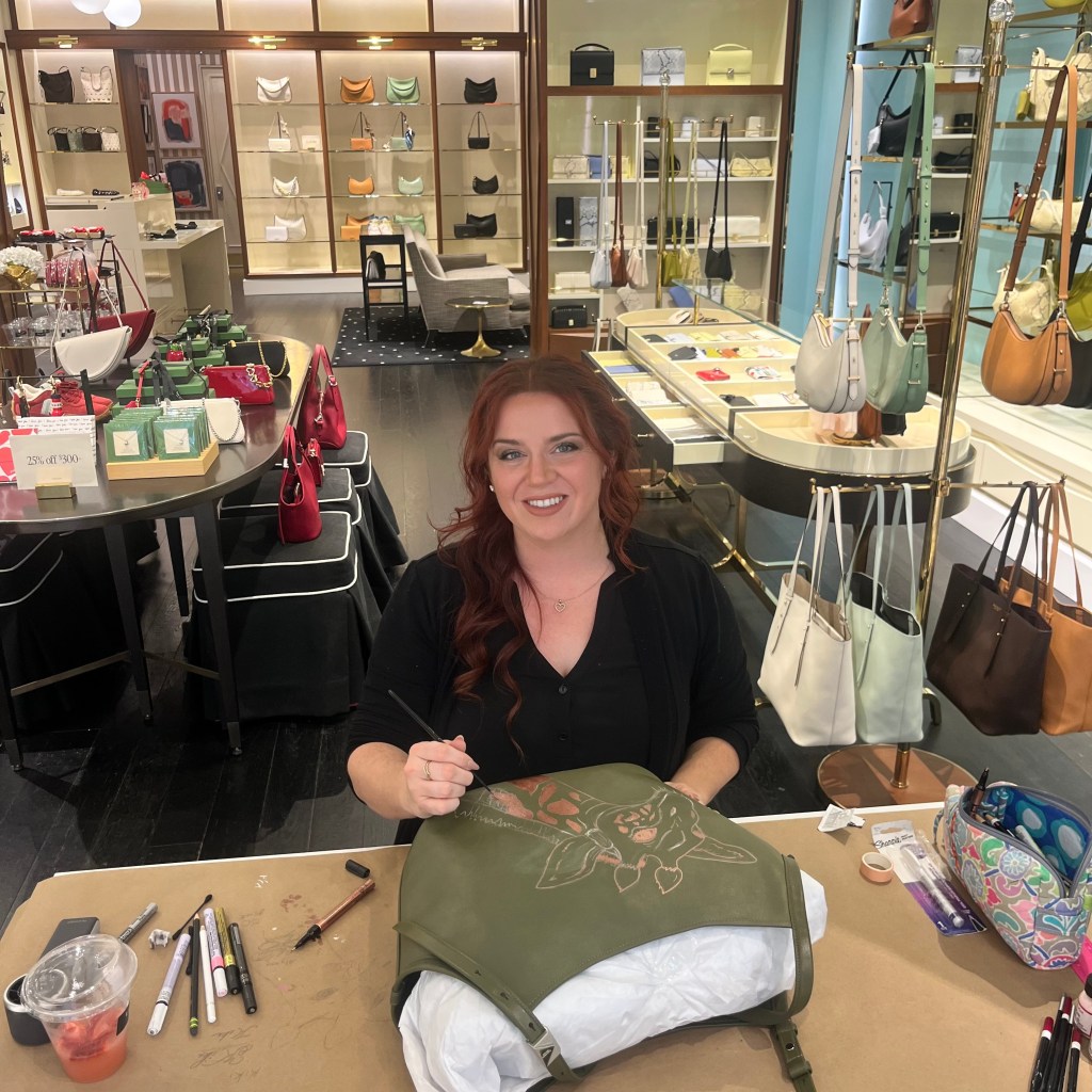 Houston calligrapher hand painting a copper metallic giraffe illustration on a green Kate Spade handbag during a live luxury personalization event.