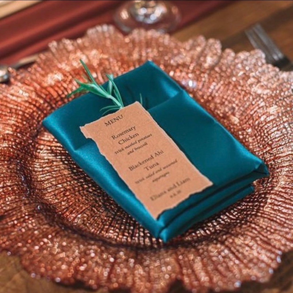 Hand torn wedding menu with mixed typography placed on teal napkin and copper charger plate showing luxury wedding table design.