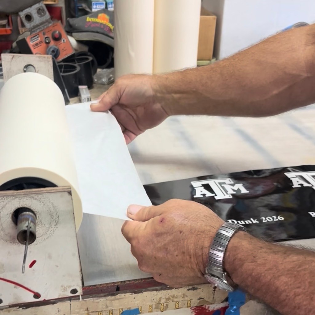 Hands applying transfer masking to a custom vinyl stencil in a sign shop, preparing a Texas A&M logo design for glass etching.