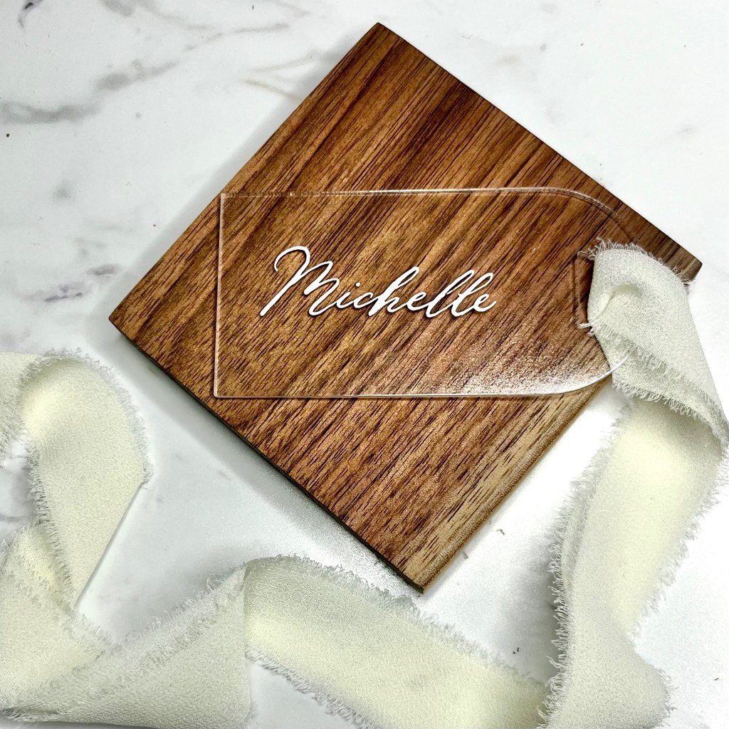 Custom acrylic bridesmaid gift tag for Michelle with white calligraphy lettering and soft yellow ribbon displayed on a walnut wood coaster.