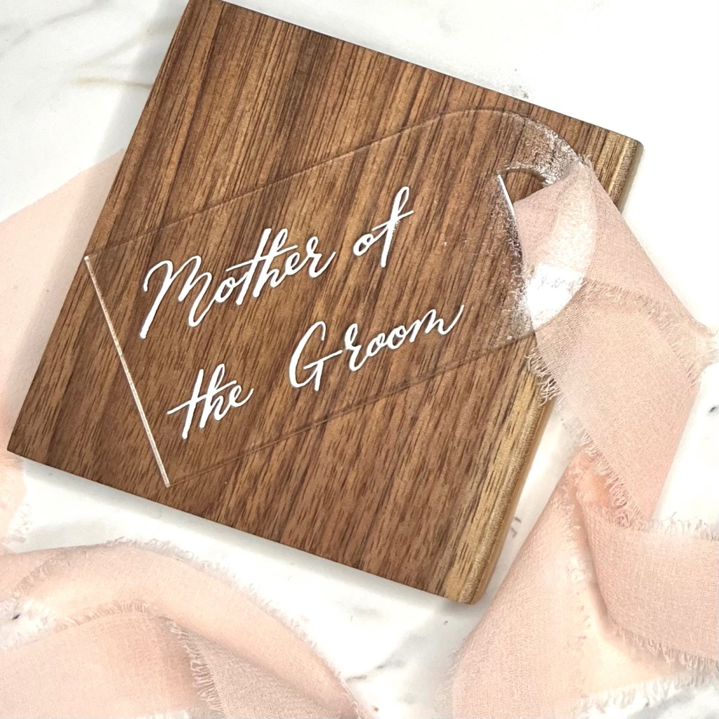 Finished Mother of the Groom acrylic wedding gift tag with white calligraphy lettering and blush ribbon displayed on a handcrafted walnut wood coaster.