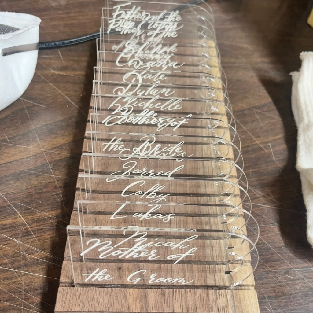 Custom acrylic wedding place cards drying in a handcrafted walnut rack after lettering and finishing, preparing for final wedding installation.