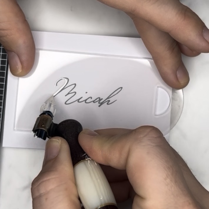 Pointed pen calligraphy being written on an acrylic wedding party gift tag during the lettering process.