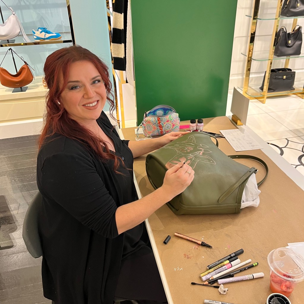 Calligraphy artist hand painting a leather handbag during a live personalization event at Kate Spade, demonstrating the craftsmanship behind custom luxury accessories.